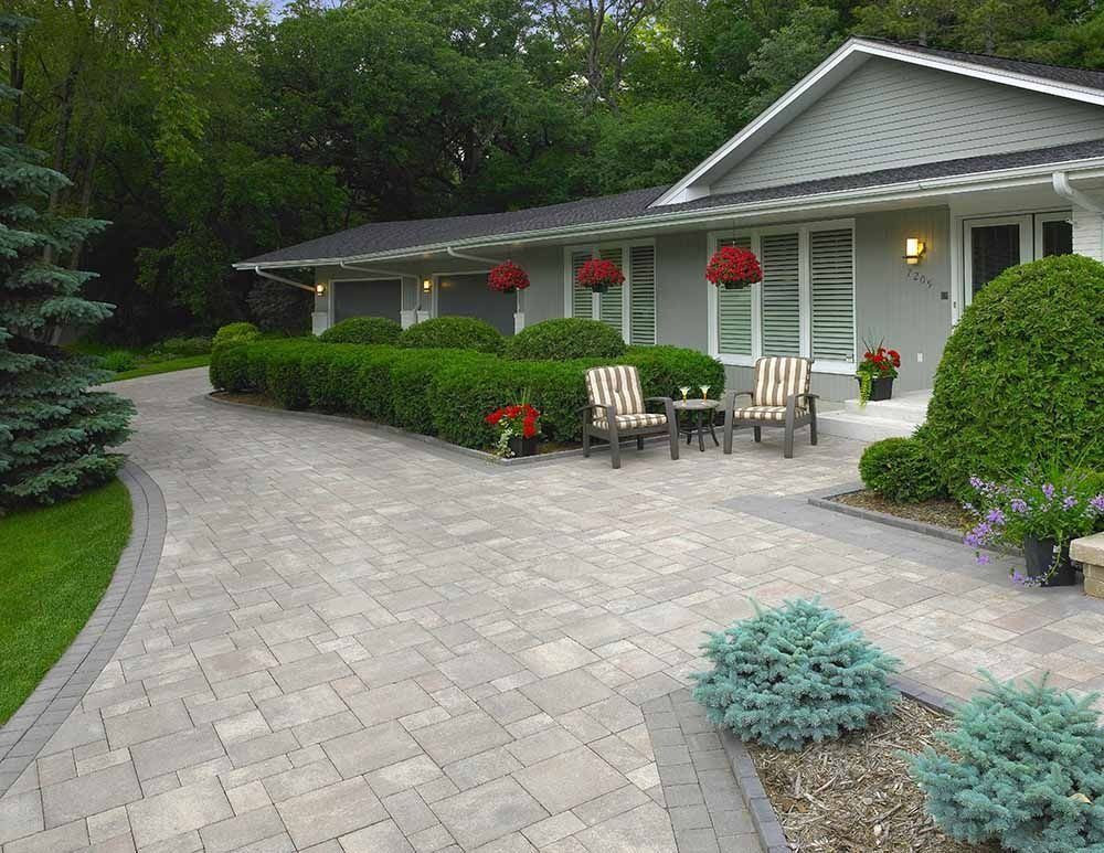 A gray ranch-style house with a light gray paver driveway, manicured hedges, and two patio chairs near the front entrance.