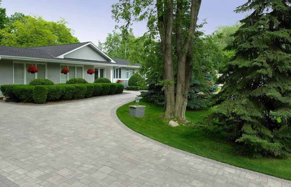 A light grey house with a dark roof and a curved stone driveway, surrounded by green lawns and trees.