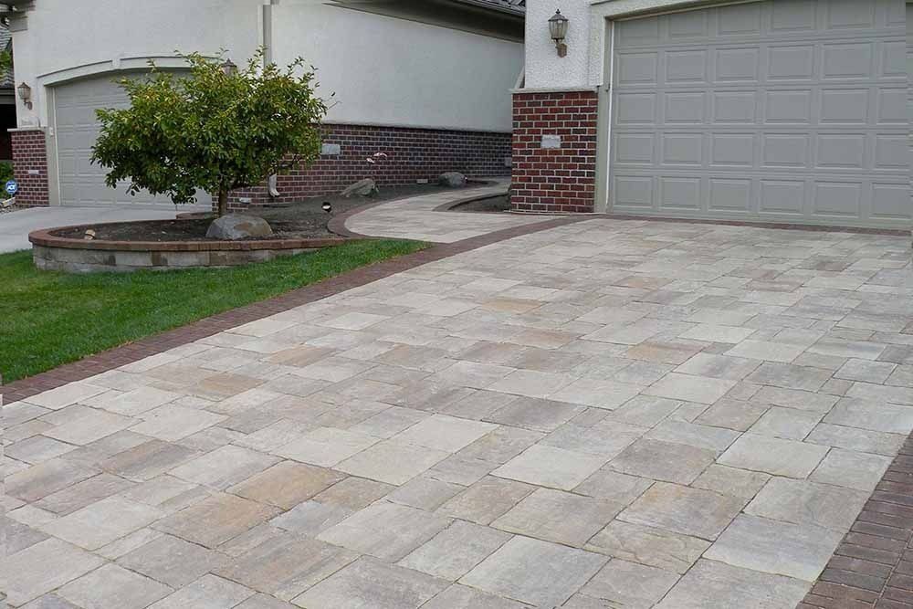 A paver driveway in a neutral, multi-tonal gray and tan pattern leading to a brick-accented house with a garage.