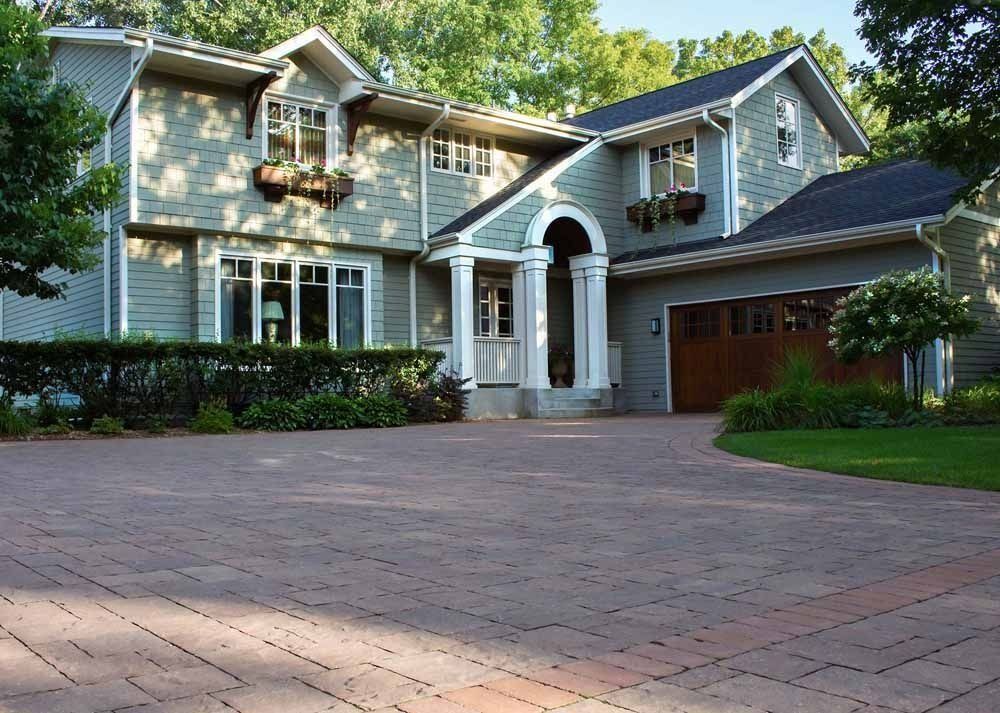A two-story green house with a white-pillared entrance and a red brick paver driveway.
