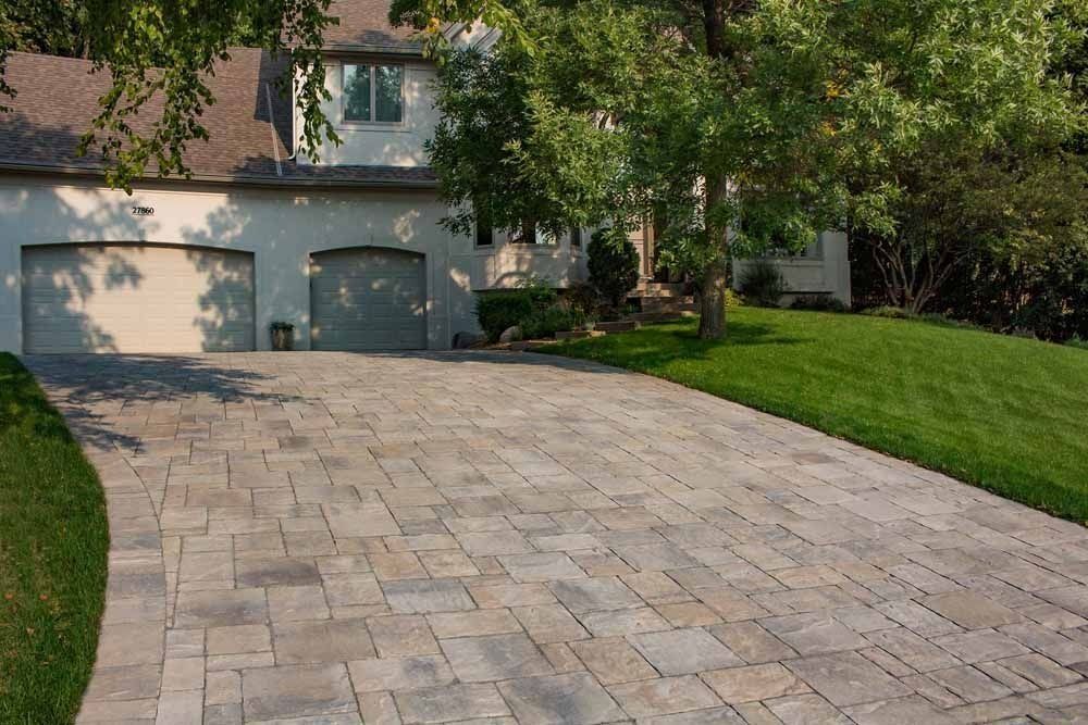 A paved stone driveway leads to a two-car garage of a white house surrounded by green trees and lawn.