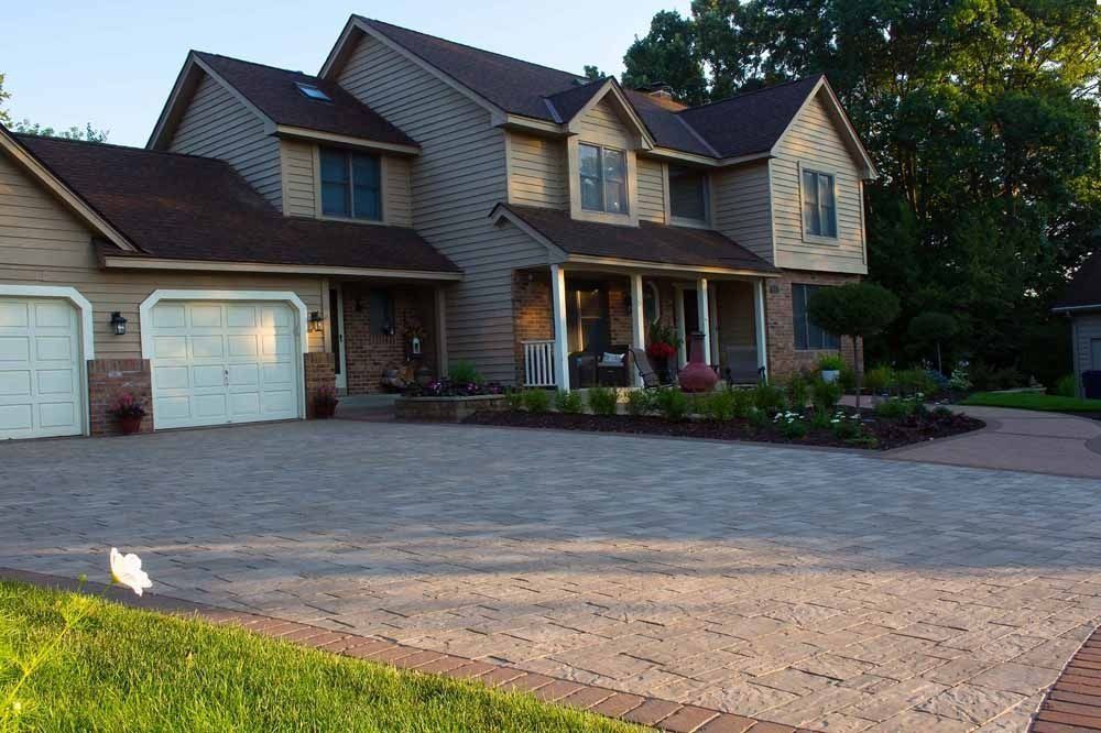 A large two-story suburban home with tan siding, a dark roof, and a wide, paved driveway with a red brick border.