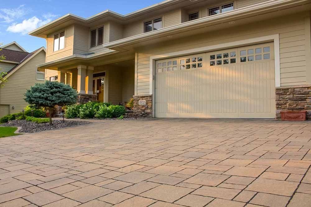 A two-story suburban home with tan siding, a stone facade, and a paved brick driveway leading to a large garage.