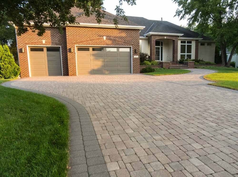 A tan paver driveway with a dark border leads to a brick house with a two-car garage and a front entrance.