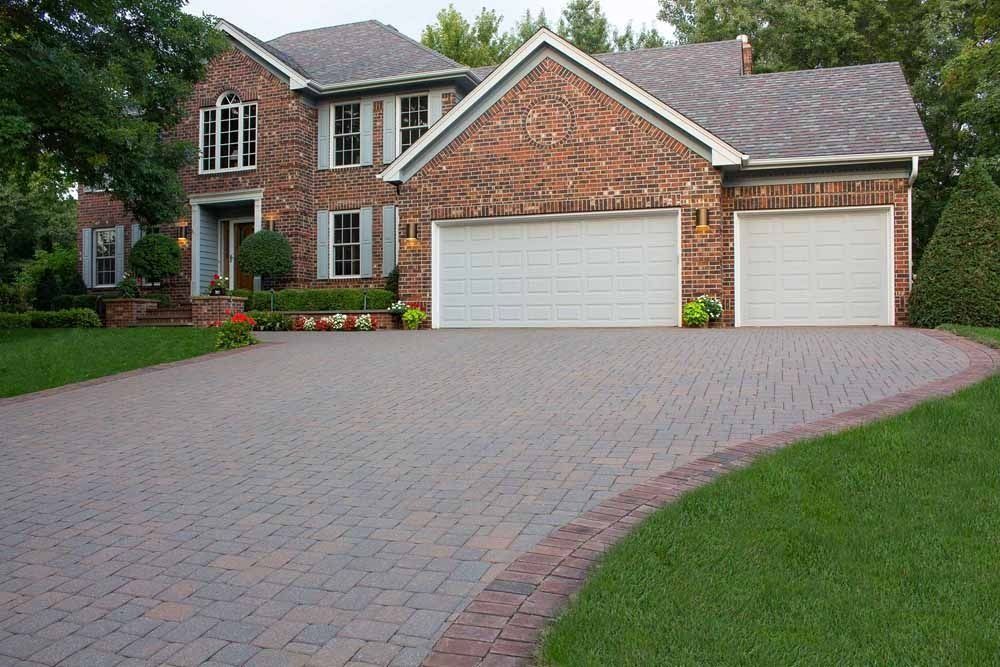 A large two-story brick house with a paved driveway and a three-car garage, surrounded by a lawn and green trees.