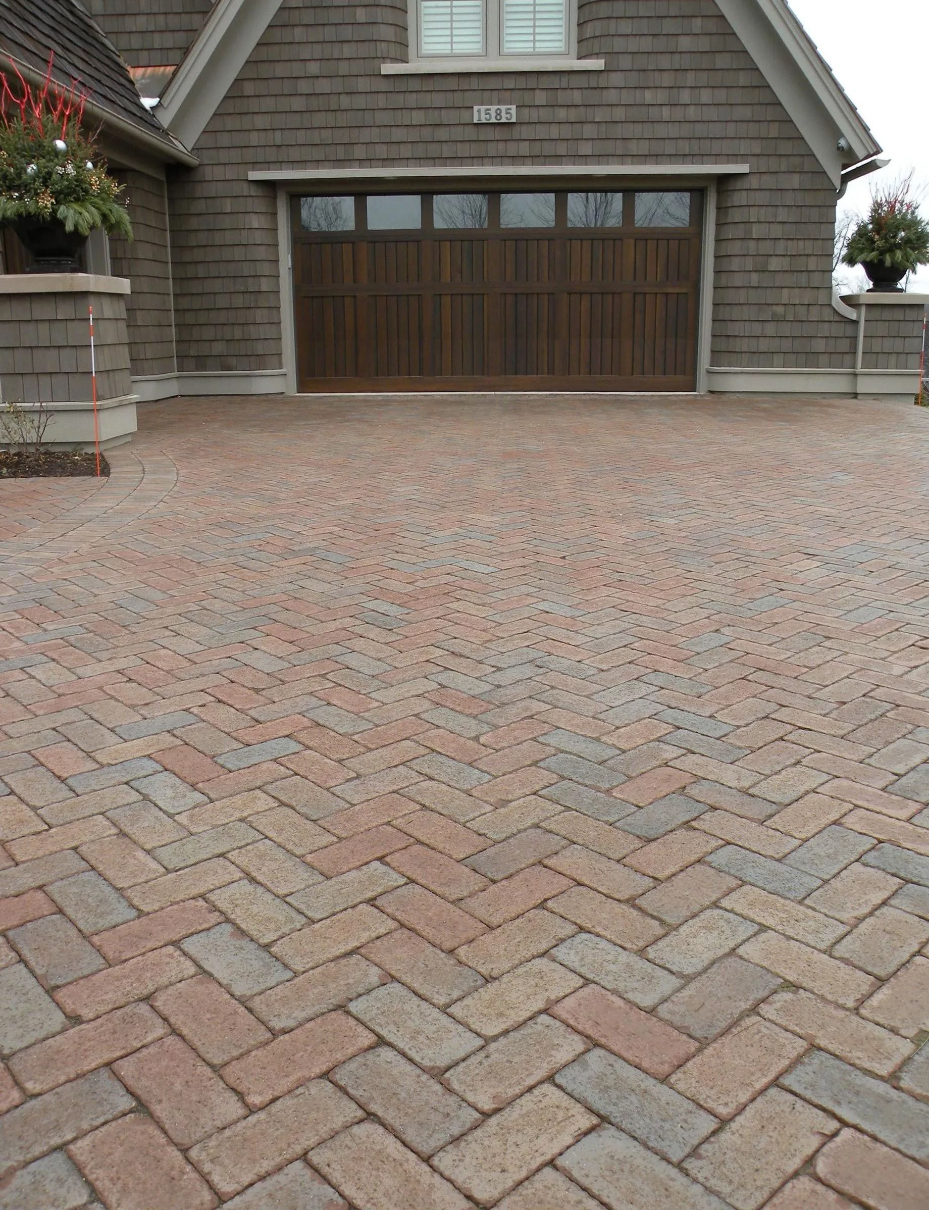 A multi-colored brick driveway in a herringbone pattern leads to a wooden garage door on a brown shingled house.