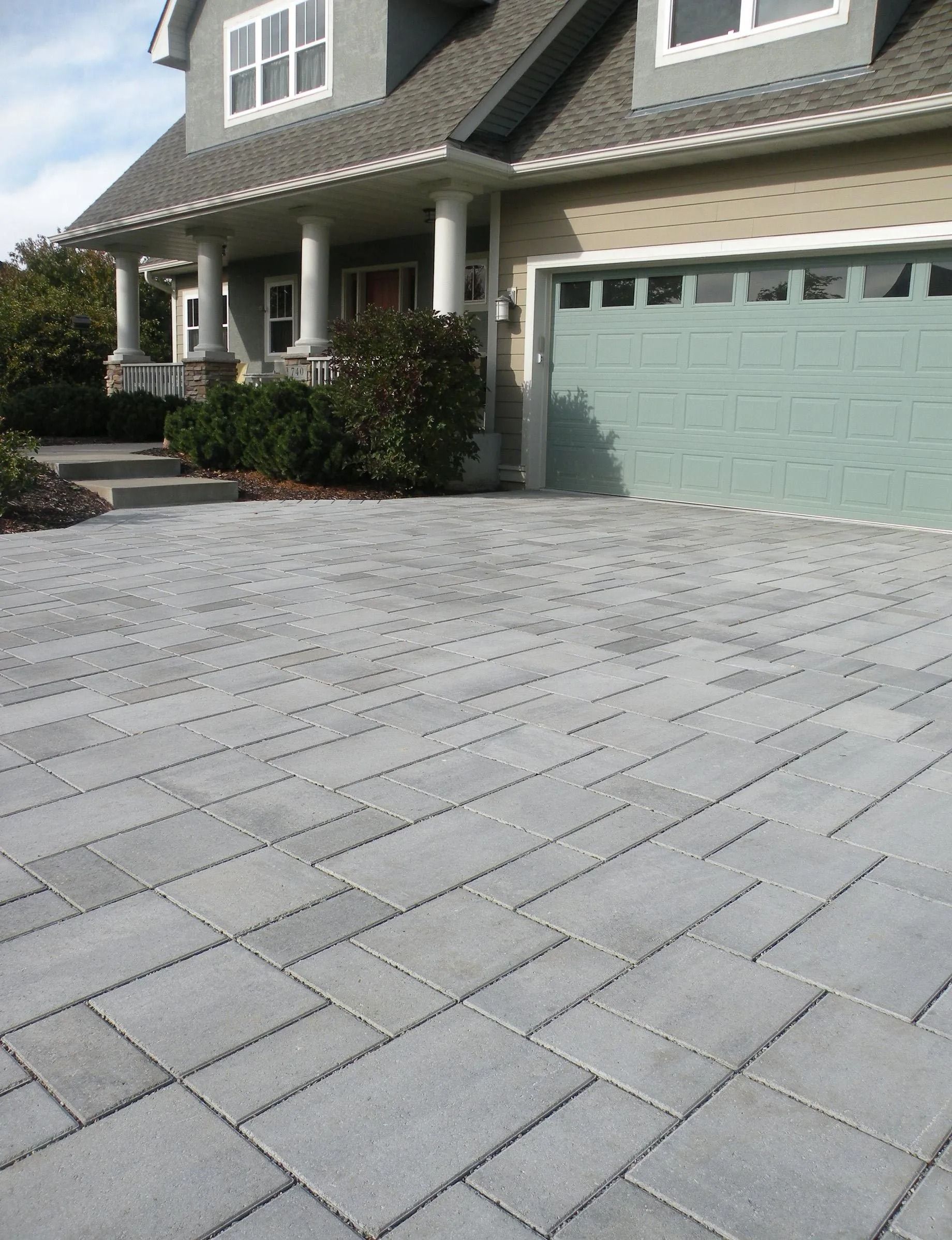A paved driveway with a rectangular gray paver pattern leading to a house with a green garage door and front columns.