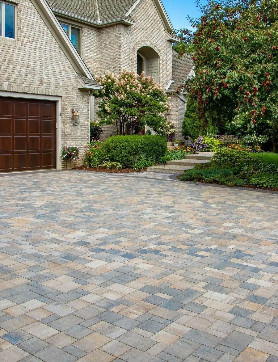 A stone-paved driveway leads to a house with a brown garage door, surrounded by lush green trees and landscaping.