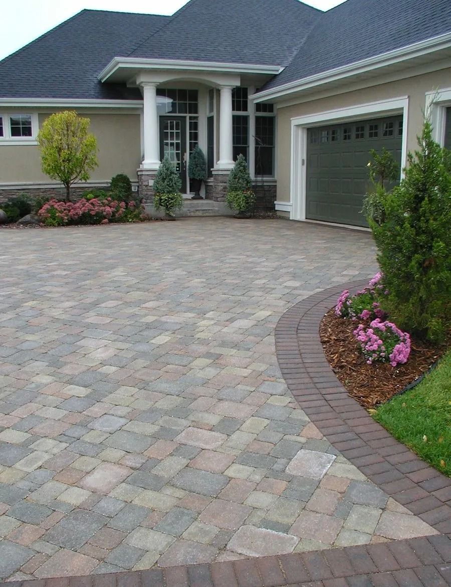 A driveway paved with multi-colored stone pavers with a darker red brick border leading to a suburban home garage.