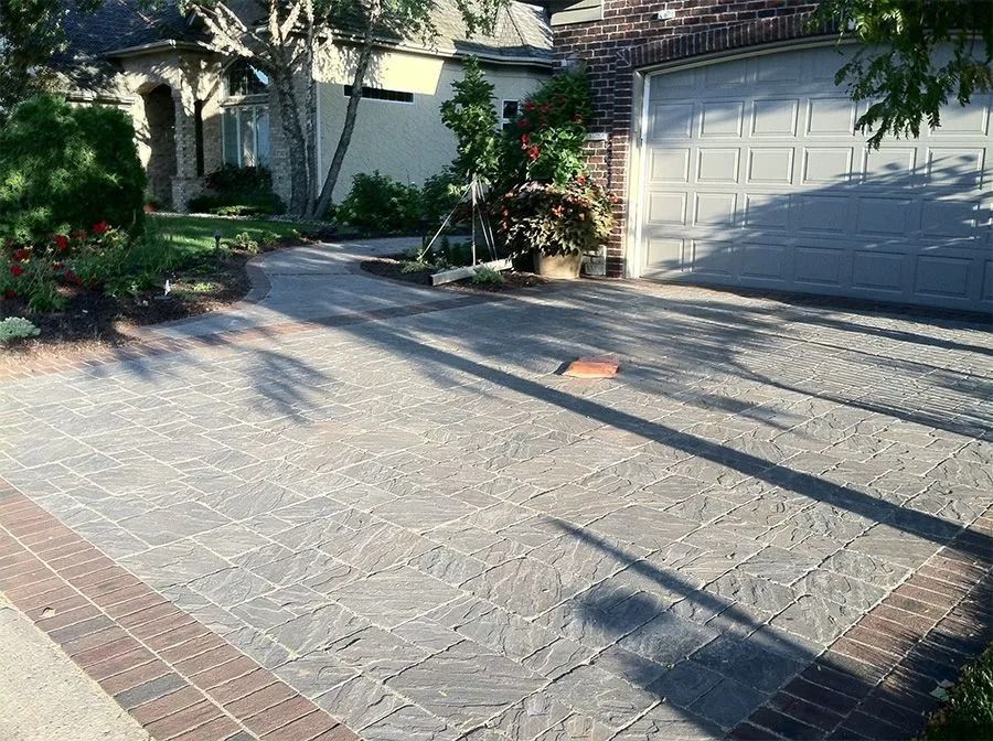 A residential driveway and walkway paved with grey textured stone tiles, bordered by a decorative red brick edge.