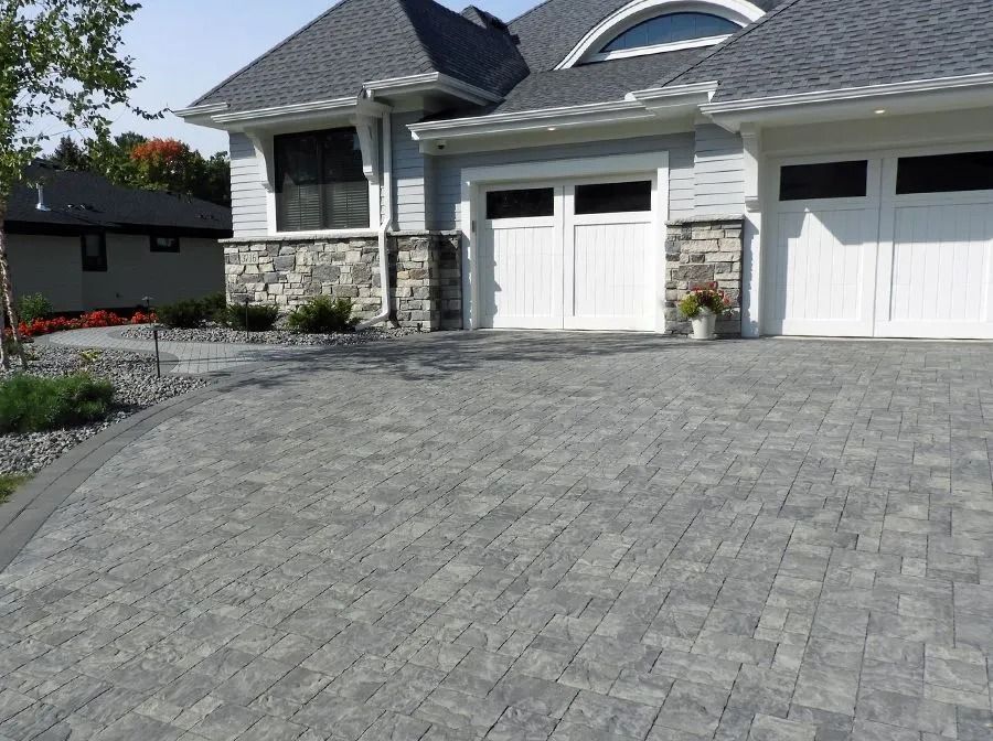 A grey paved driveway leads to a house with white garage doors and a stone-accented exterior on a sunny day.