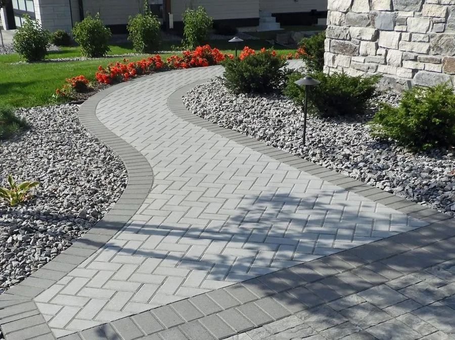 A curved paver walkway with a gray brick border winds through a landscape of decorative rocks, shrubs, and red flowers.