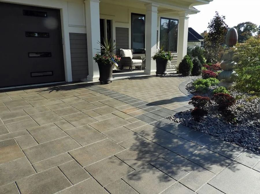 A paved stone driveway leads to a front porch and entryway of a suburban house, framed by planters and landscaping.