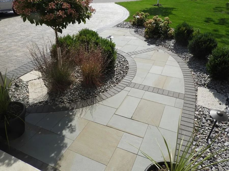 A curved stone walkway with a decorative brick border, set amidst a landscaped garden with shrubs and gray stone gravel.