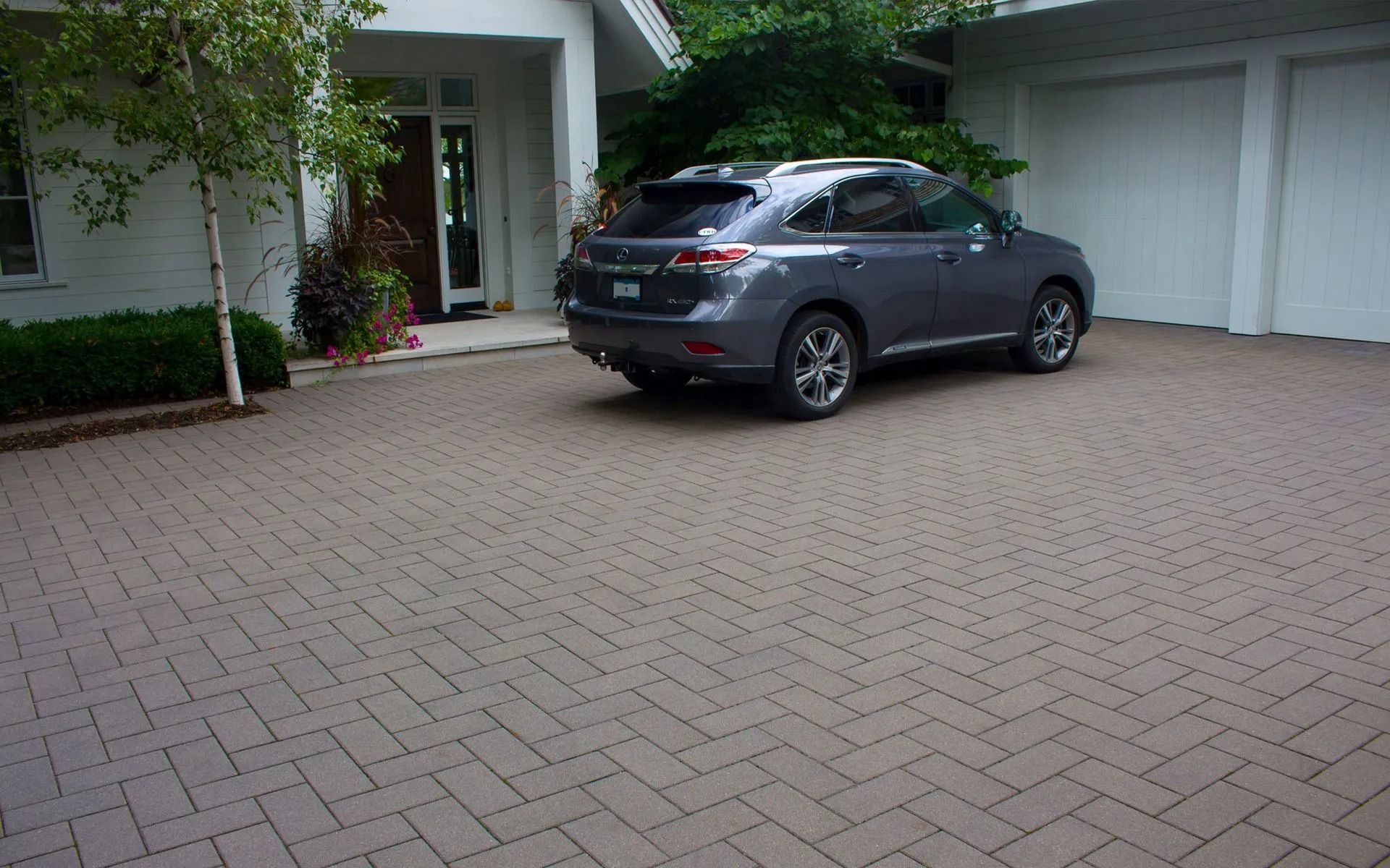 A dark gray SUV parked on a light-colored herringbone-patterned paver driveway in front of a house.