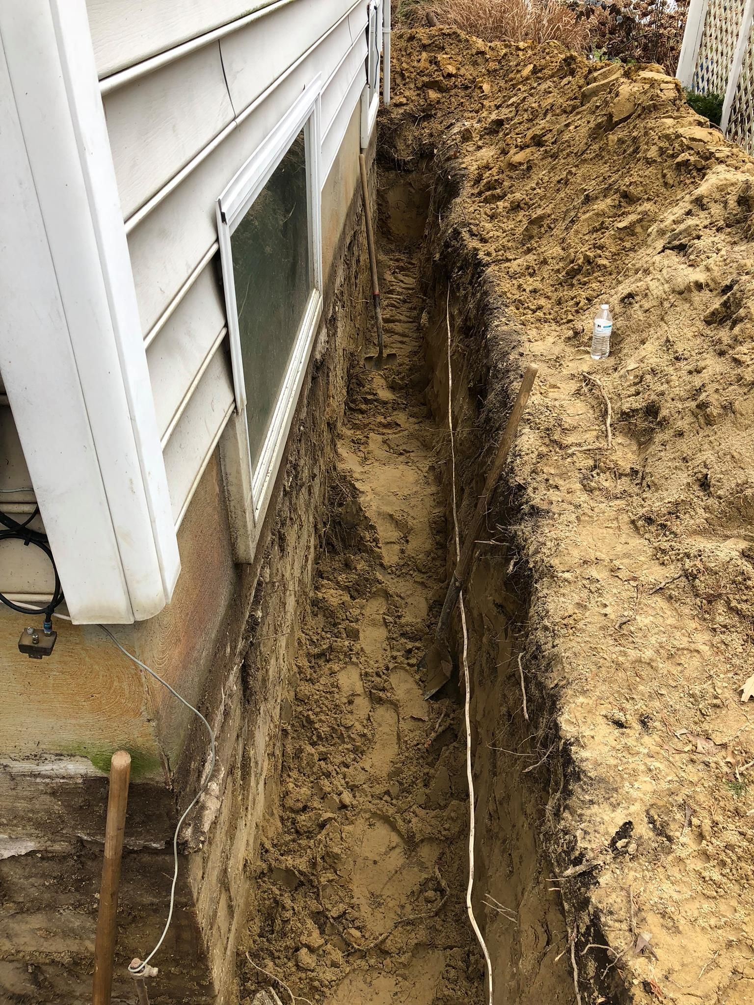 Narrow trench dug beside a house foundation, with exposed soil and siding visible.
