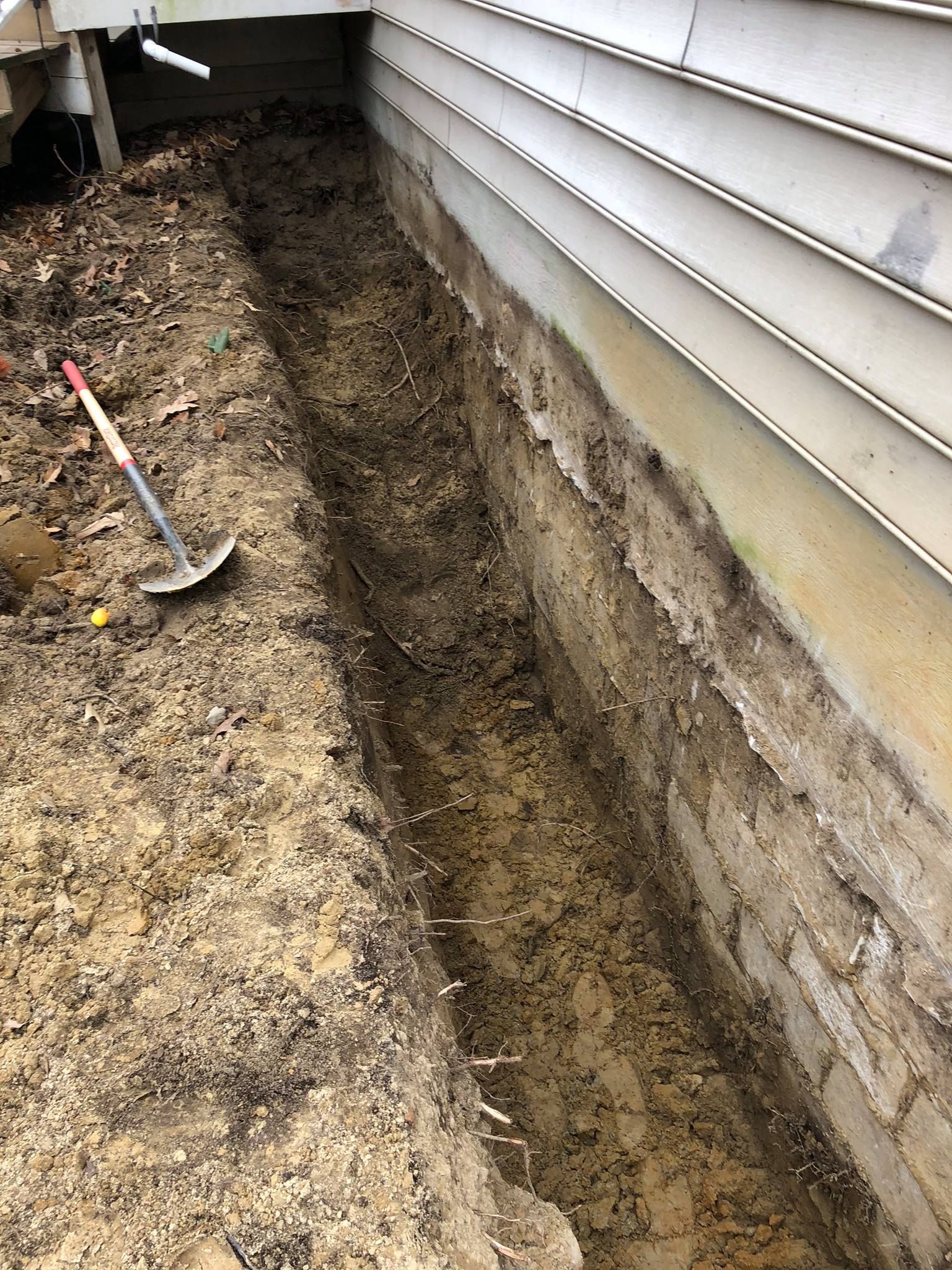 Narrow trench dug beside a house foundation, with a shovel standing in the dirt.