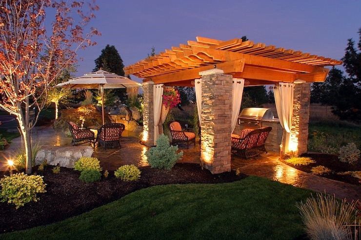 Illuminated garden gazebo with pergola roof and curtains at dusk, surrounded by landscaped greenery