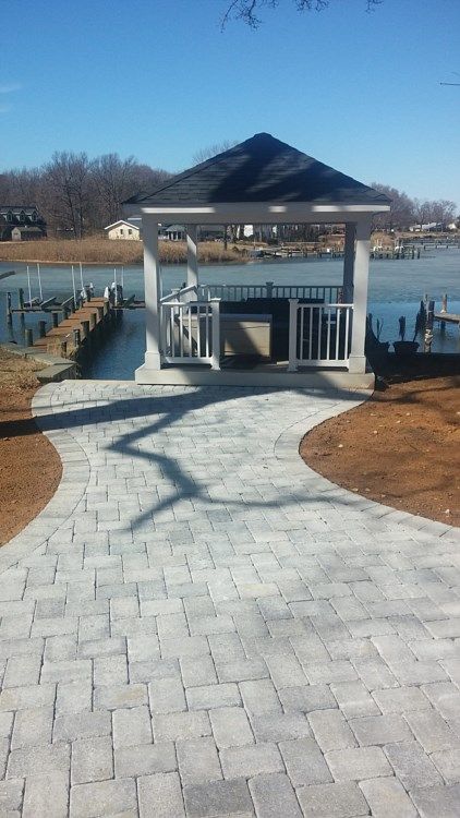 White gazebo at the end of a stone path by a calm lakeside in winter, with patches of snow.