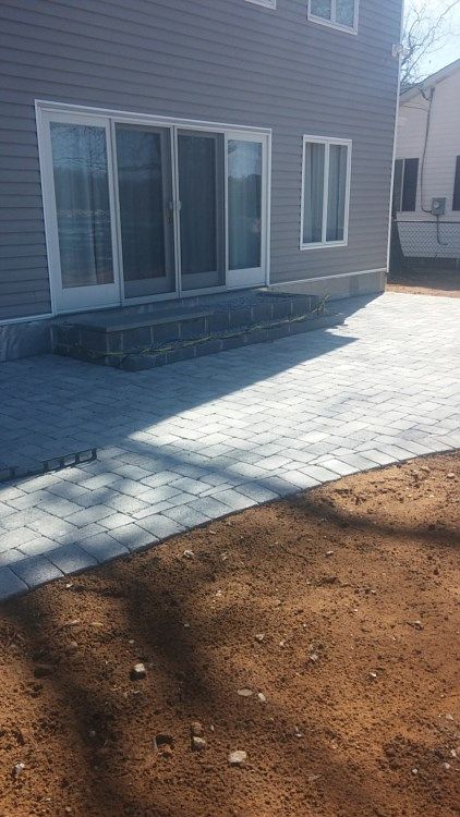 Backyard patio with gray pavers beside a house, bordered by bare soil and stone edging.