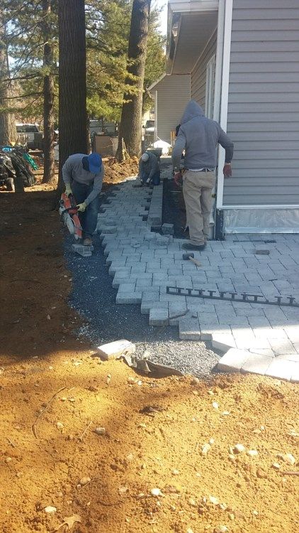Two workers laying gray pavers beside a house on a dirt path