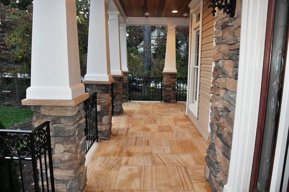 Covered front porch with stone columns, wood ceiling, and tan tiled floor overlooking greenery