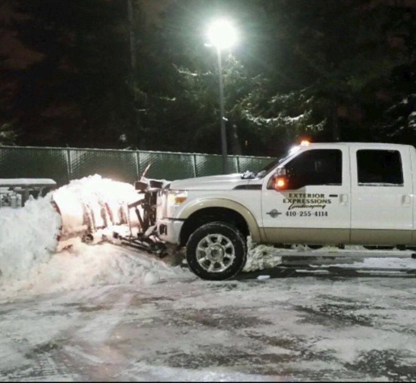 White pickup truck clearing snow with a plow in a snowy parking lot at night