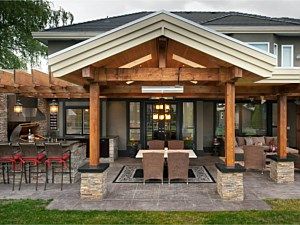 Covered patio with wood beams, outdoor dining tables, and stone fireplace in a backyard setting