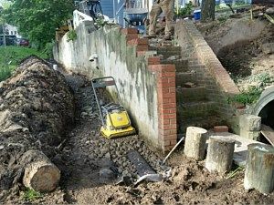 Yellow plate compactor on gravel beside a retaining wall and stairs in a worksite trench