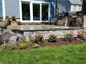 Backyard patio with stone retaining wall, glass doors, and a flower bed beside a green lawn
