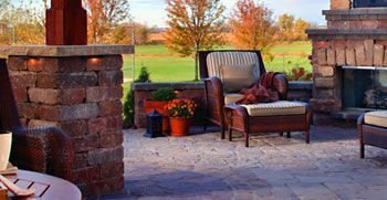 Brick patio with wicker chairs, potted flowers, and a stone fireplace in a sunny outdoor seating area
