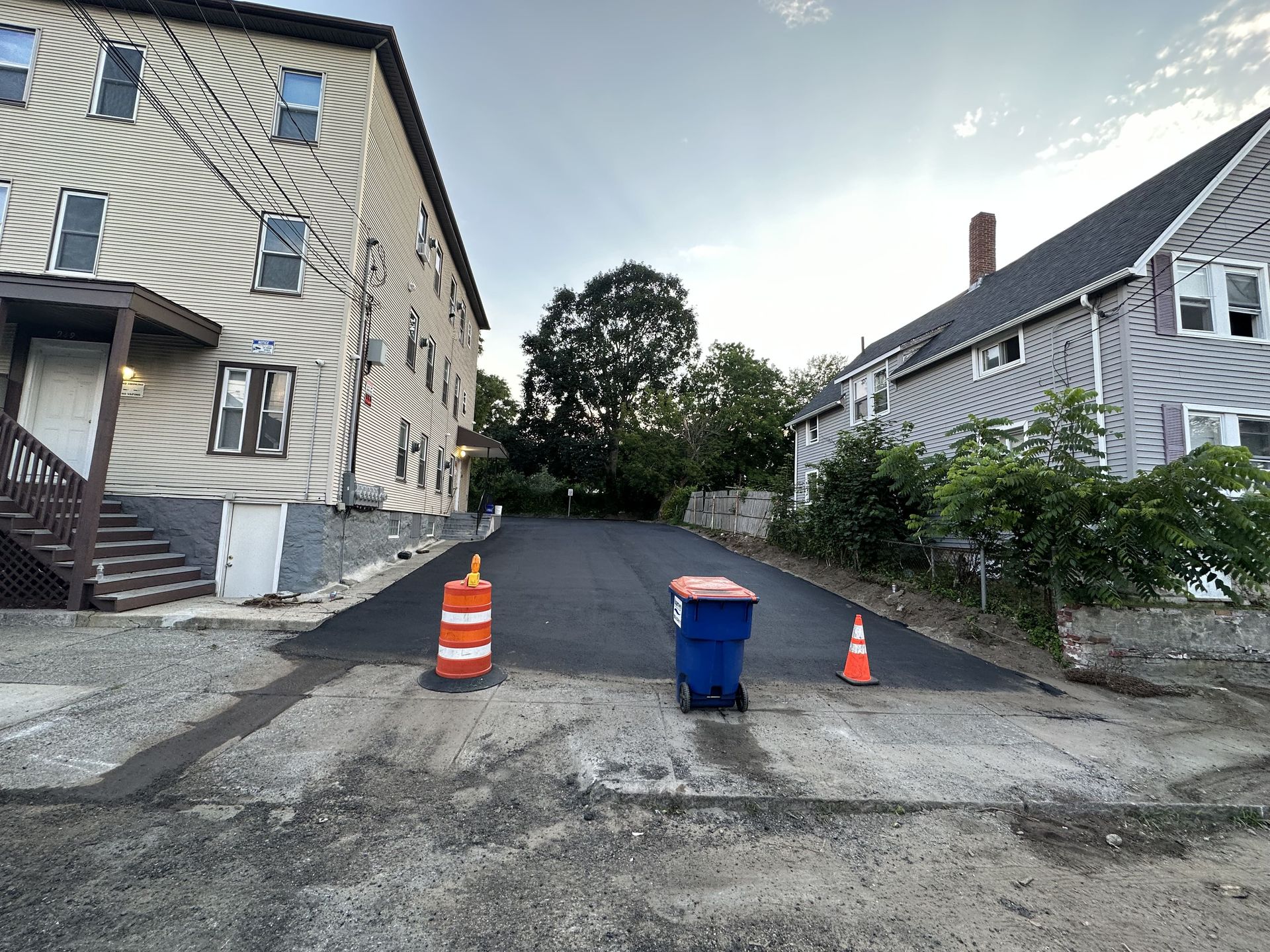 A blue trash can is sitting on the side of a road next to a building.