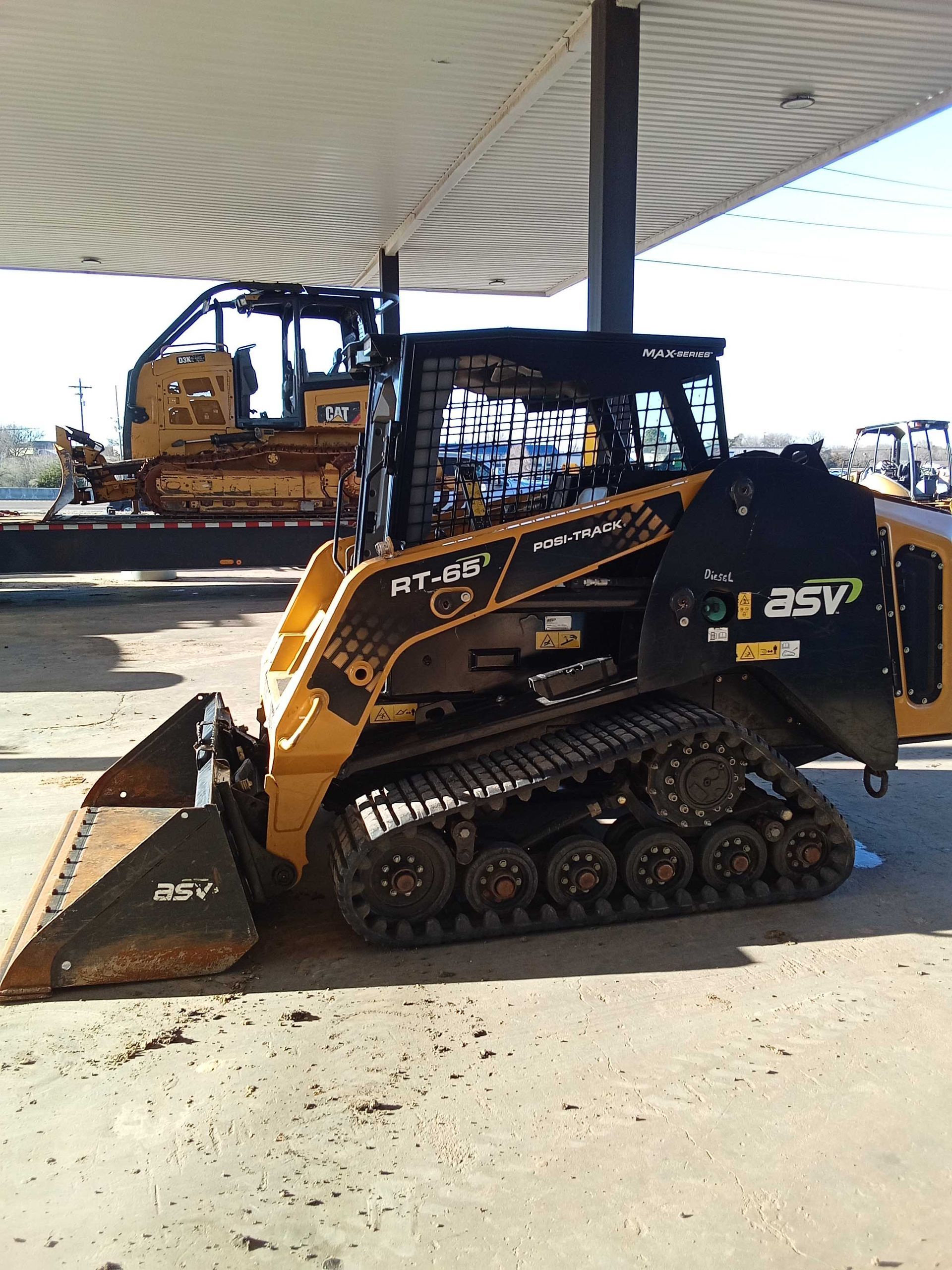 Yellow and black compact track loader parked under a canopy; bulldozer in background.