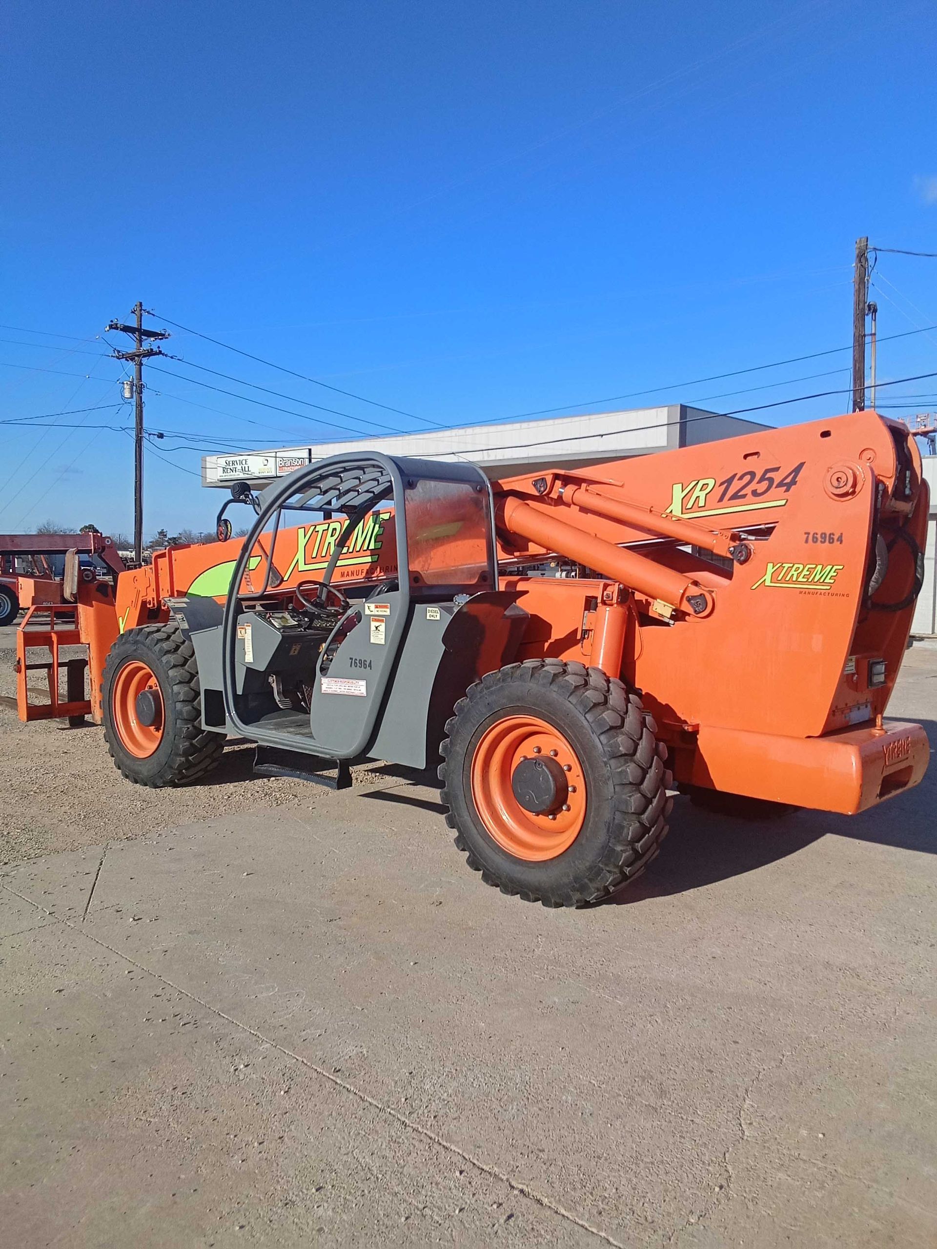 Orange telehandler on pavement under a blue sky, with utility poles and a building in the background.