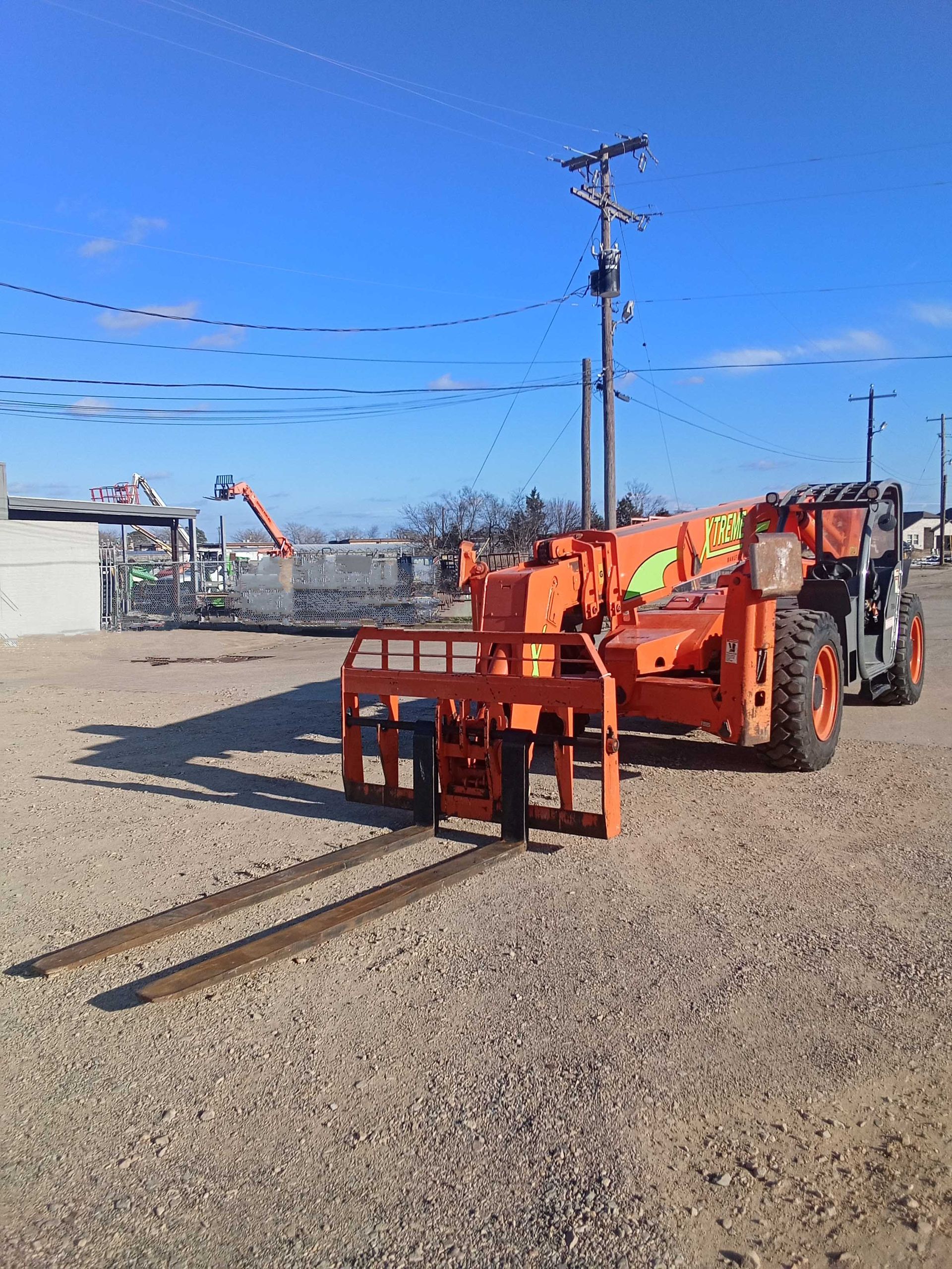 Orange forklift on gravel with extended forks under a clear blue sky.