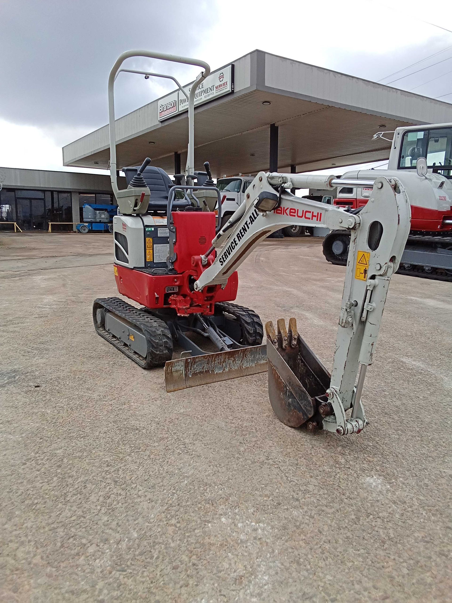 Small red and white excavator on a gravel surface, near a gas station canopy.