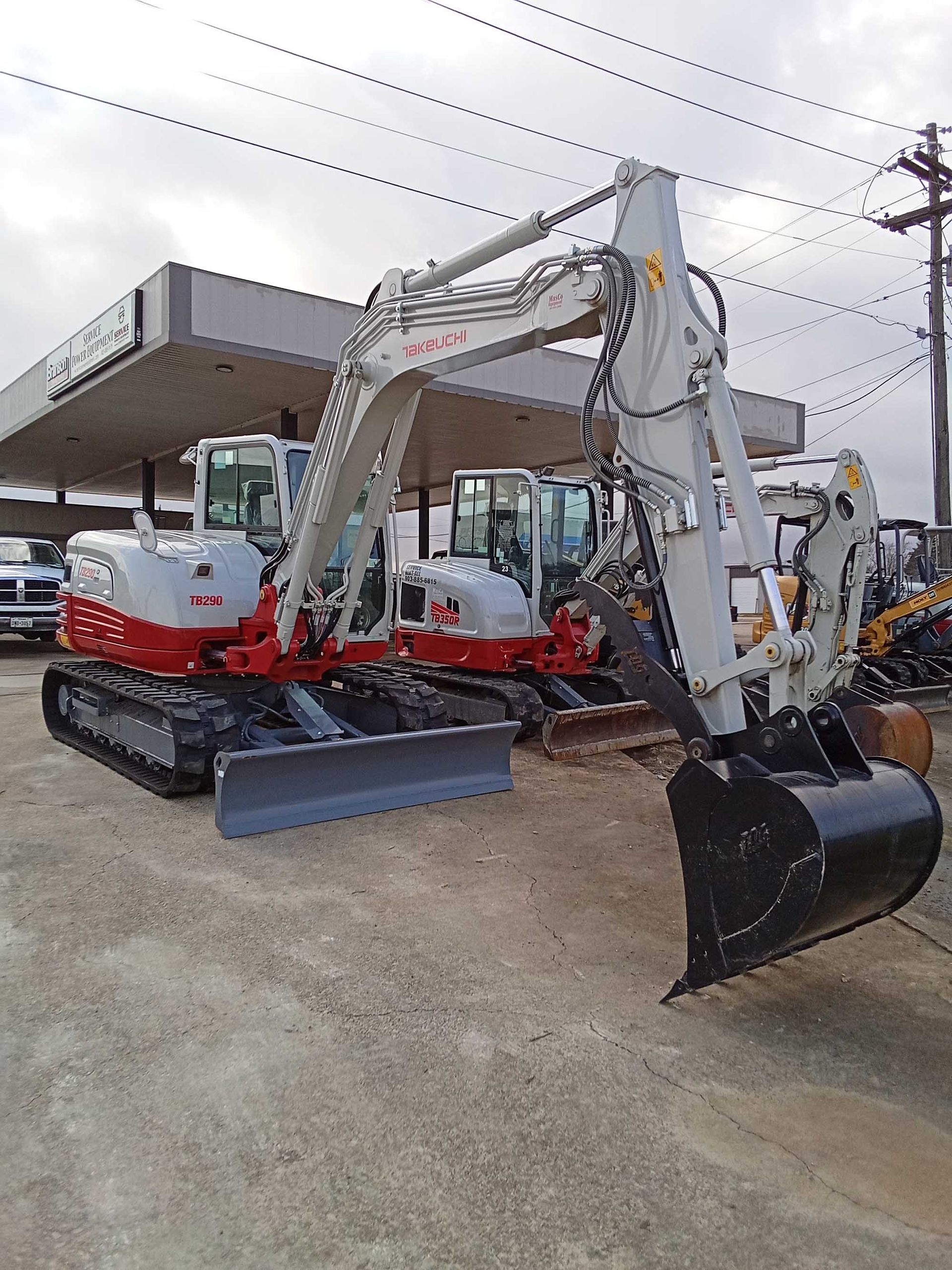 Two white and red excavators parked on a concrete lot near a gas station.