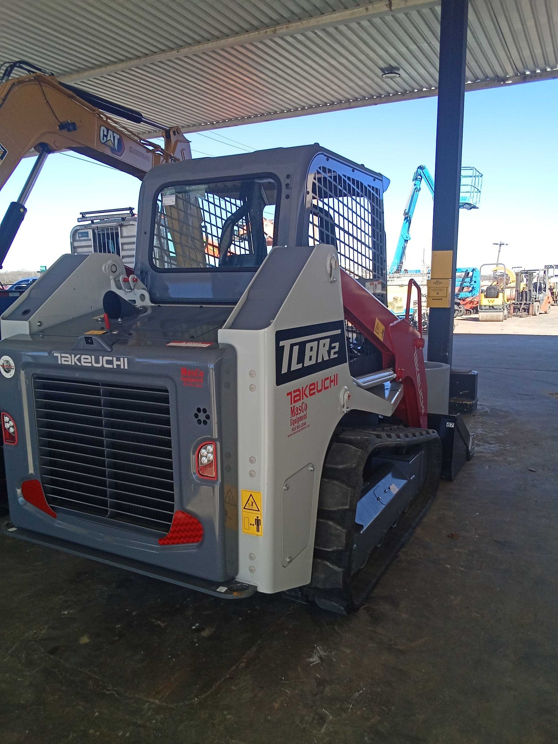 A gray and red Takeuchi TL10V2 compact track loader parked under a metal roof.