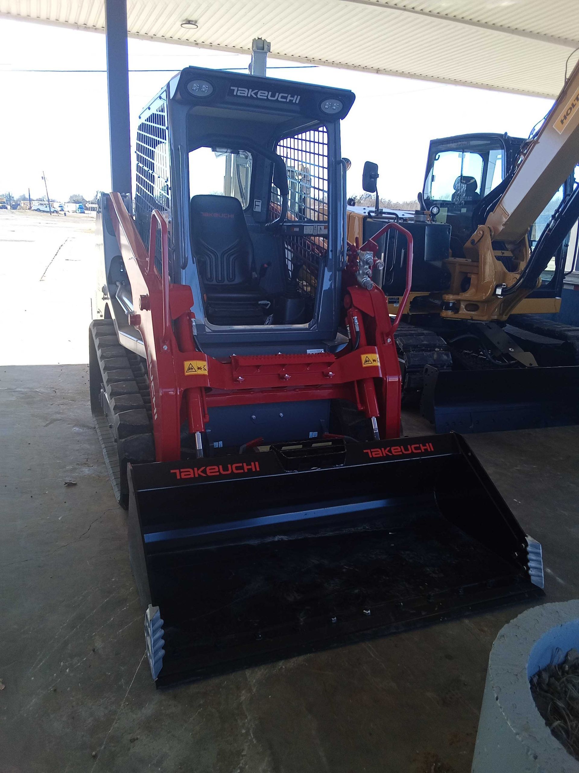Red and black compact track loader with a bucket attachment, parked under a covered structure.