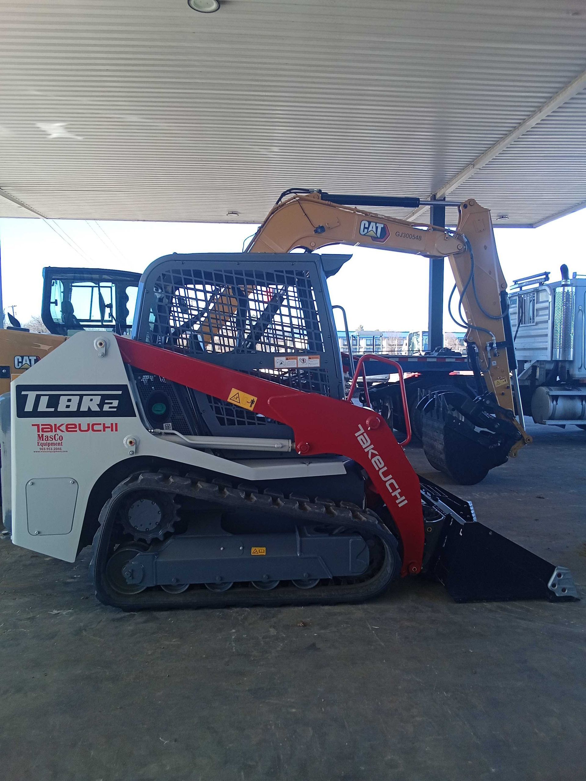 White and red track loader with black bucket parked under a canopy; yellow excavator in background.