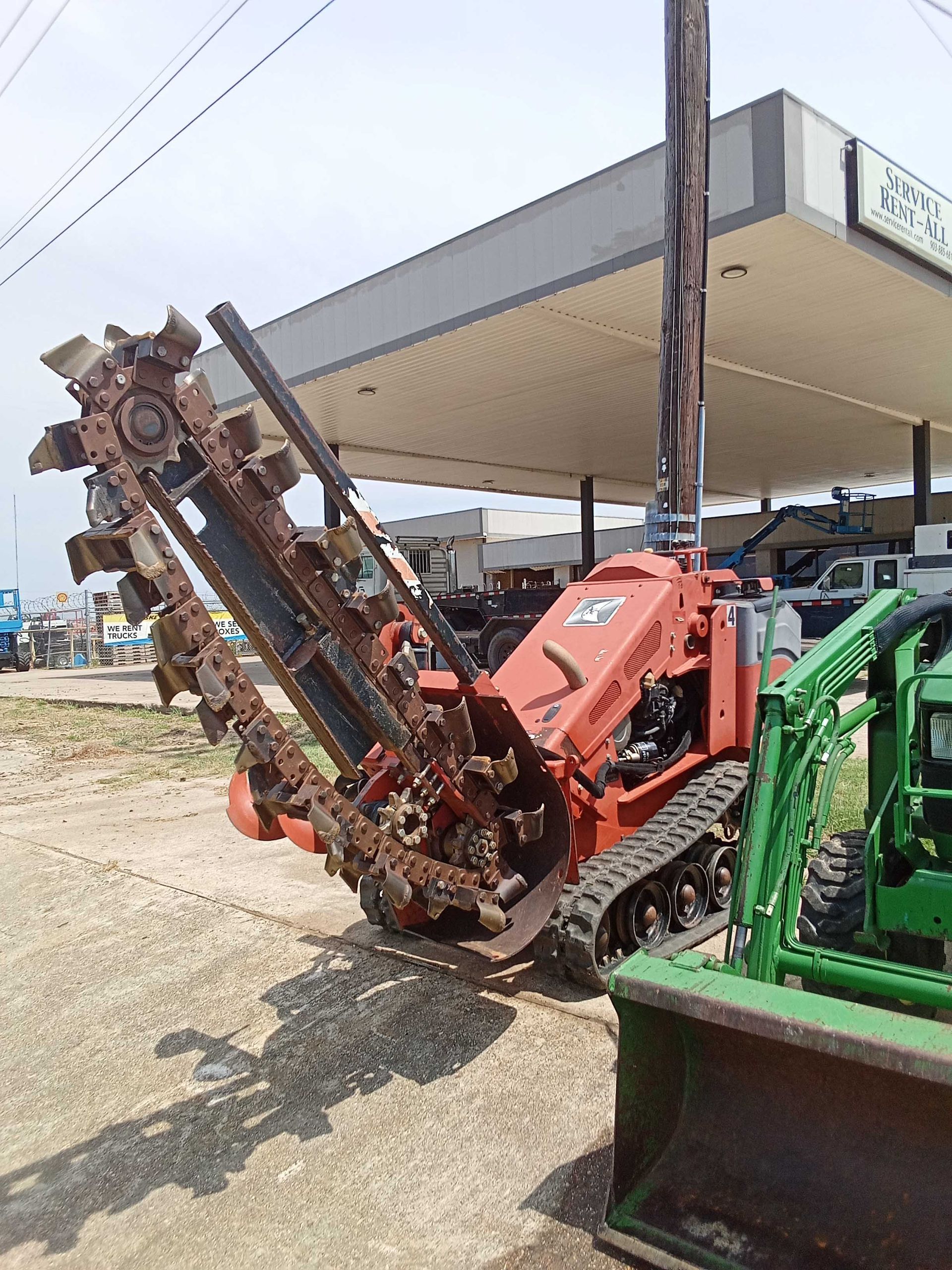 Red Ditch Witch trencher with cutting teeth, tracks, and a boom, parked next to a John Deere tractor.