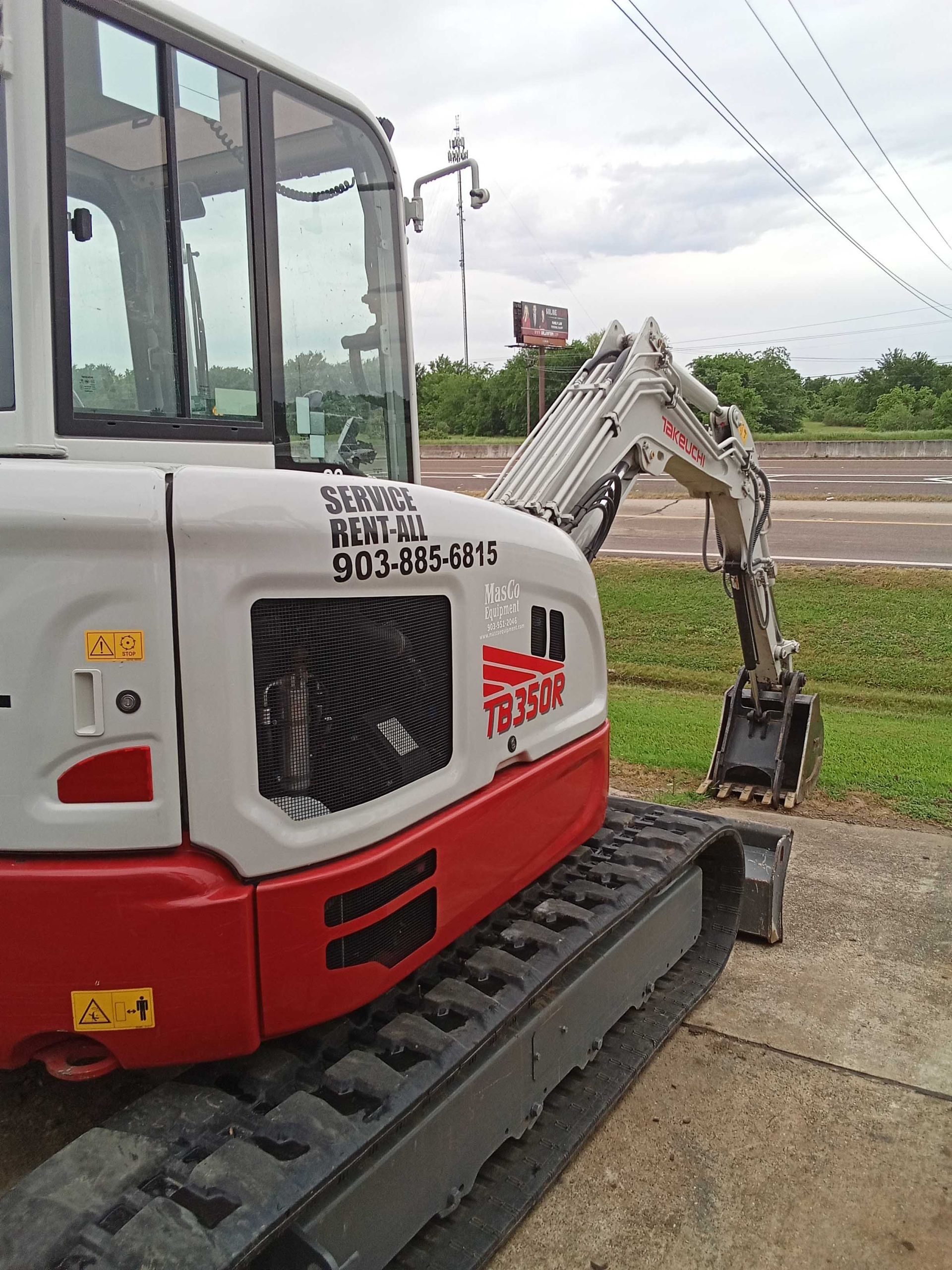 A small, red and white excavator on a paved surface near a grassy area with a cloudy sky.