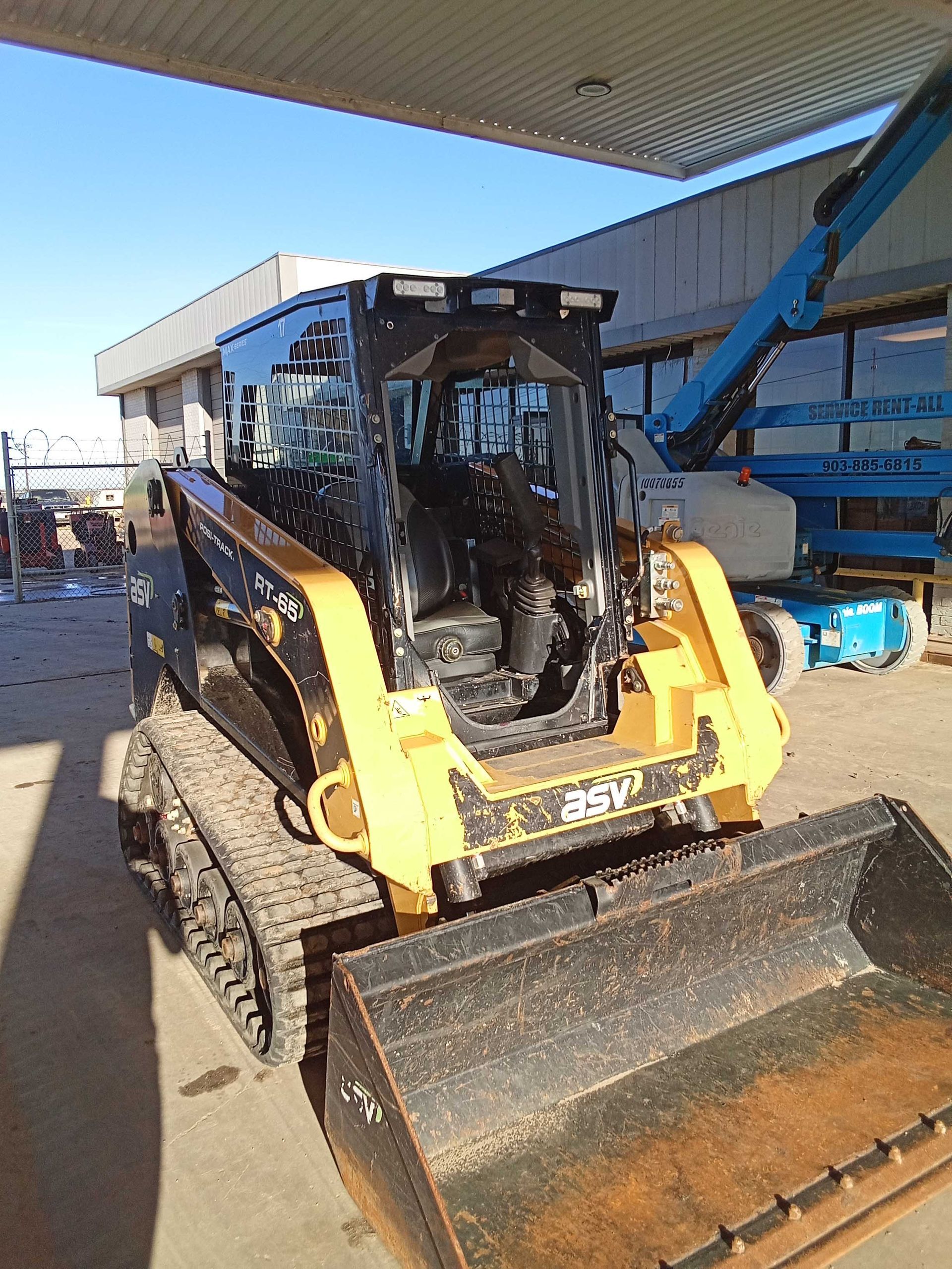 Yellow and black compact track loader with a bucket, parked near a building.