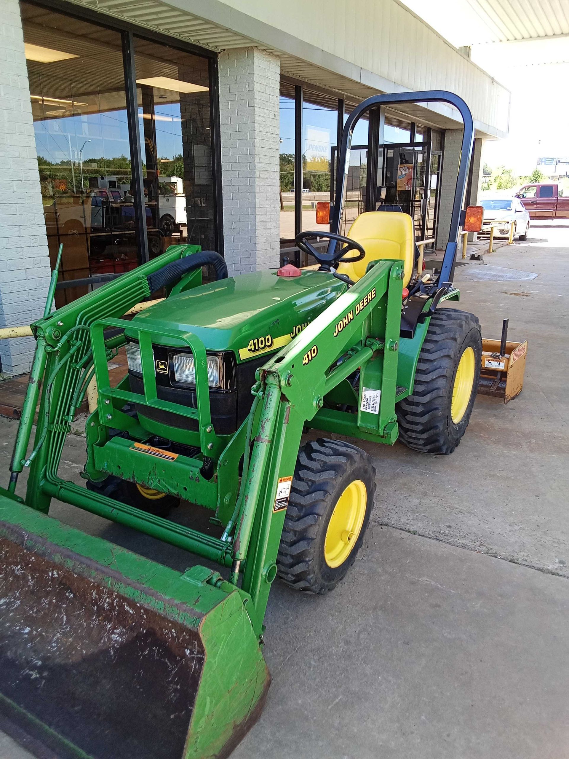 Green John Deere tractor with front loader parked outside a building with glass windows.