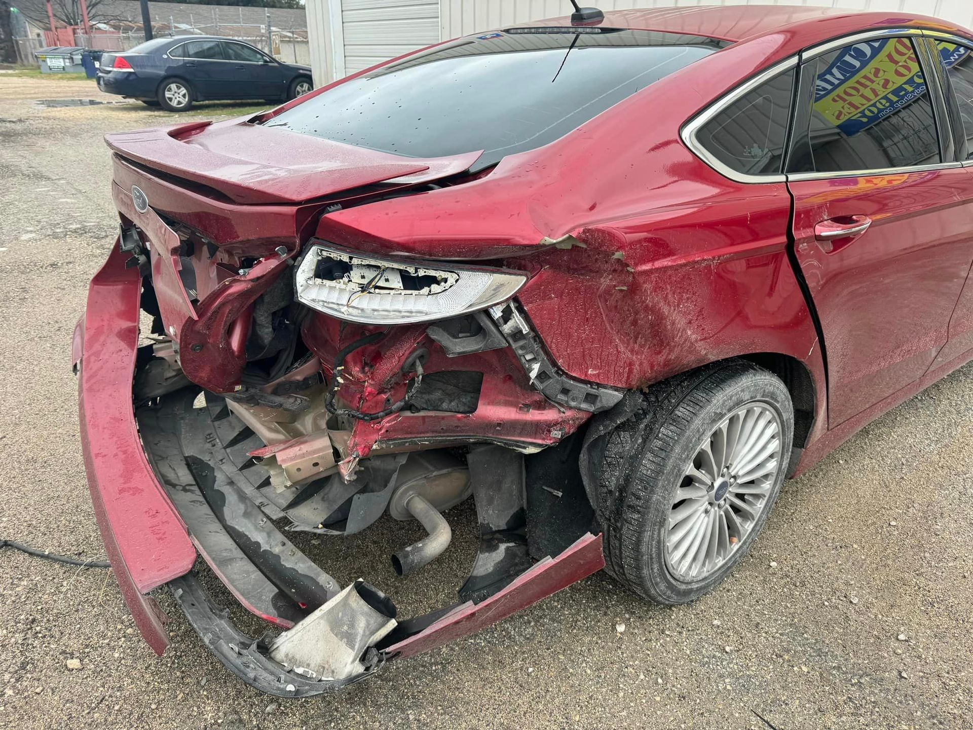A red car with a damaged front end is parked in a gravel lot.