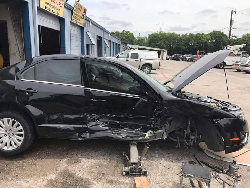 A black car with its hood open is sitting on a jack in a parking lot.