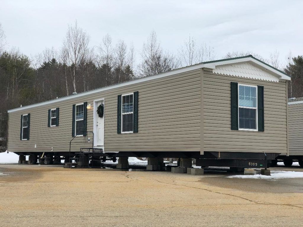 A mobile home with black shutters is parked in a gravel lot.