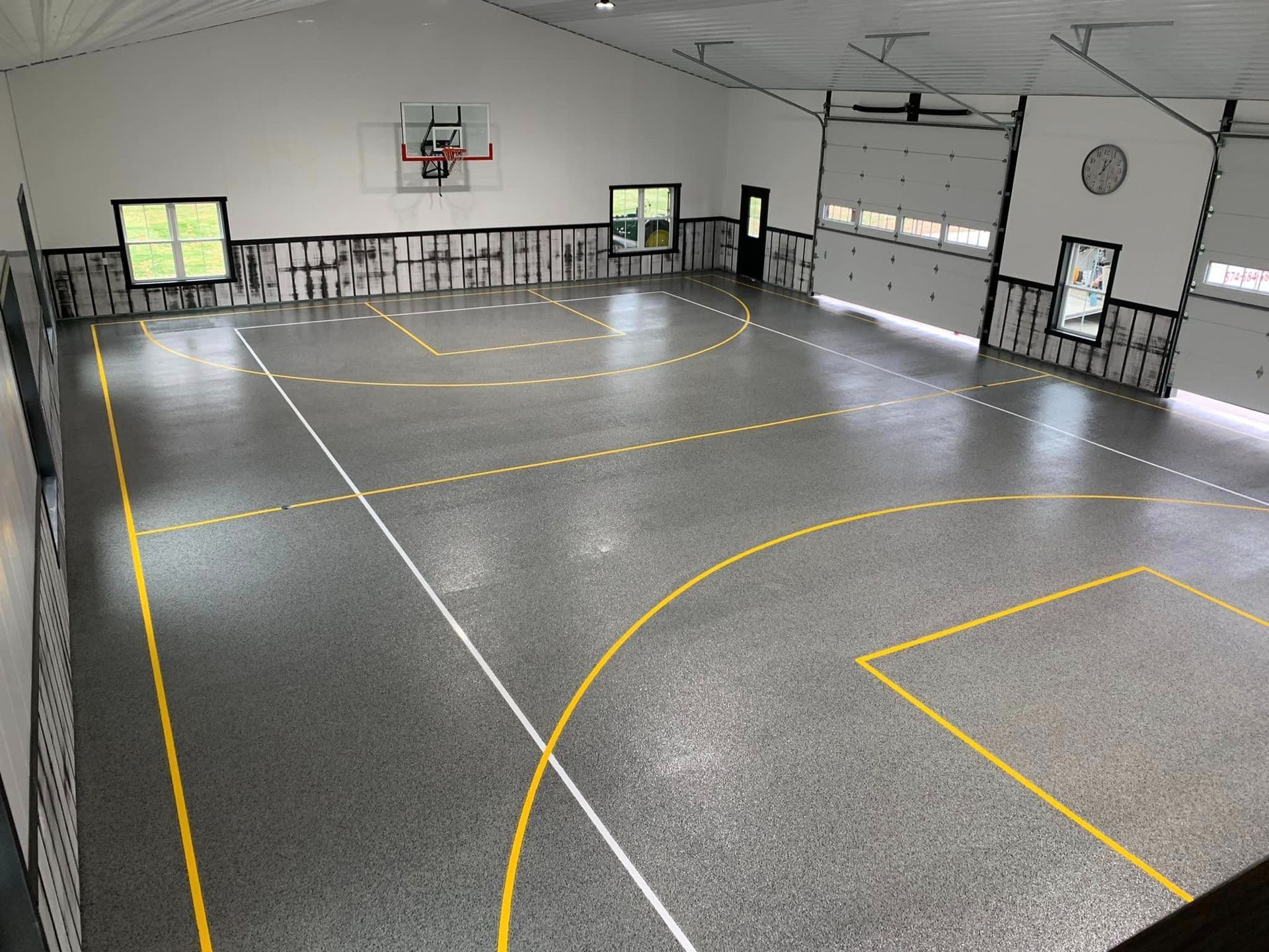 Empty indoor basketball court with gray floor, yellow lines, hoop, and large windows.