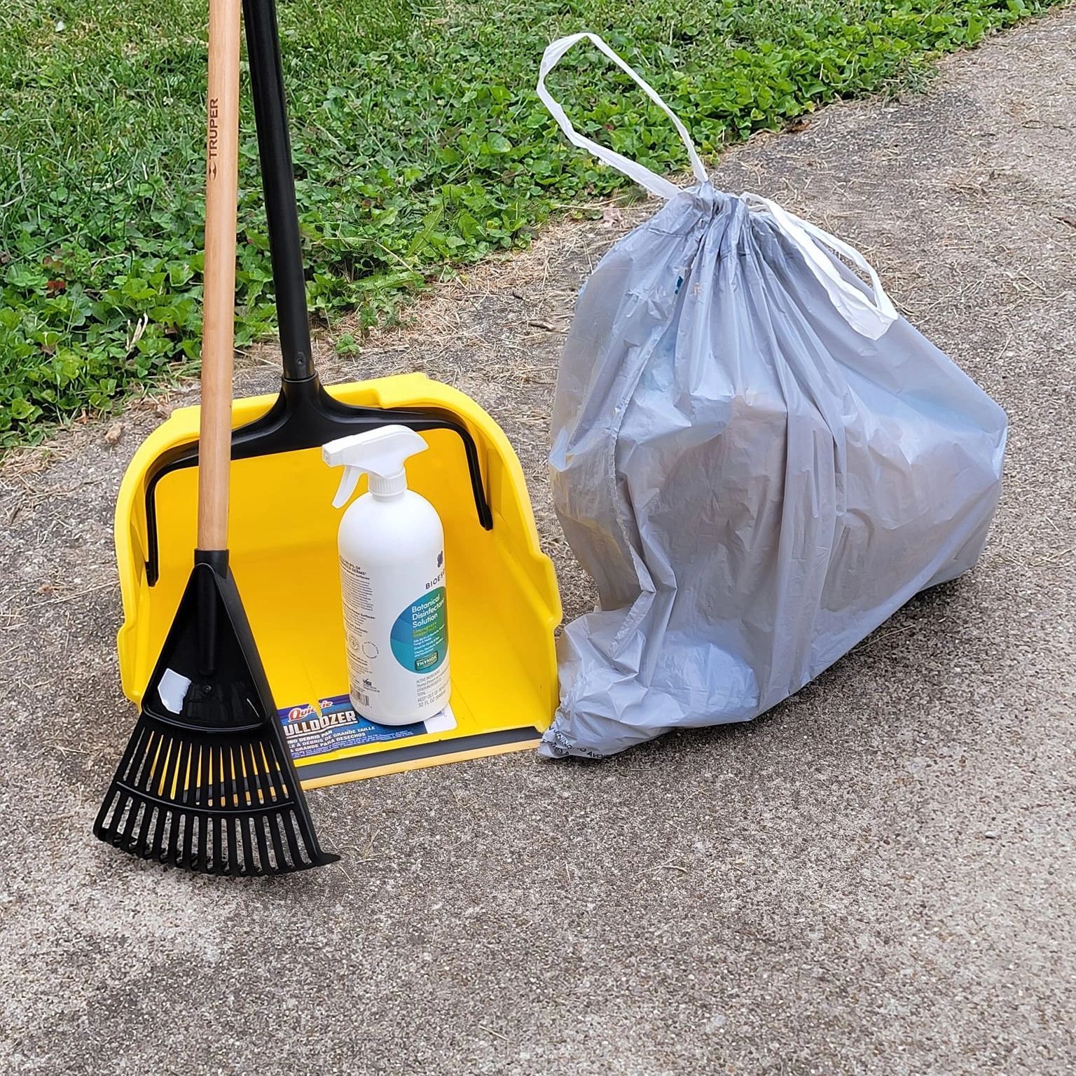 Yellow dustpan with broom, spray bottle, and gray trash bag on a sidewalk.