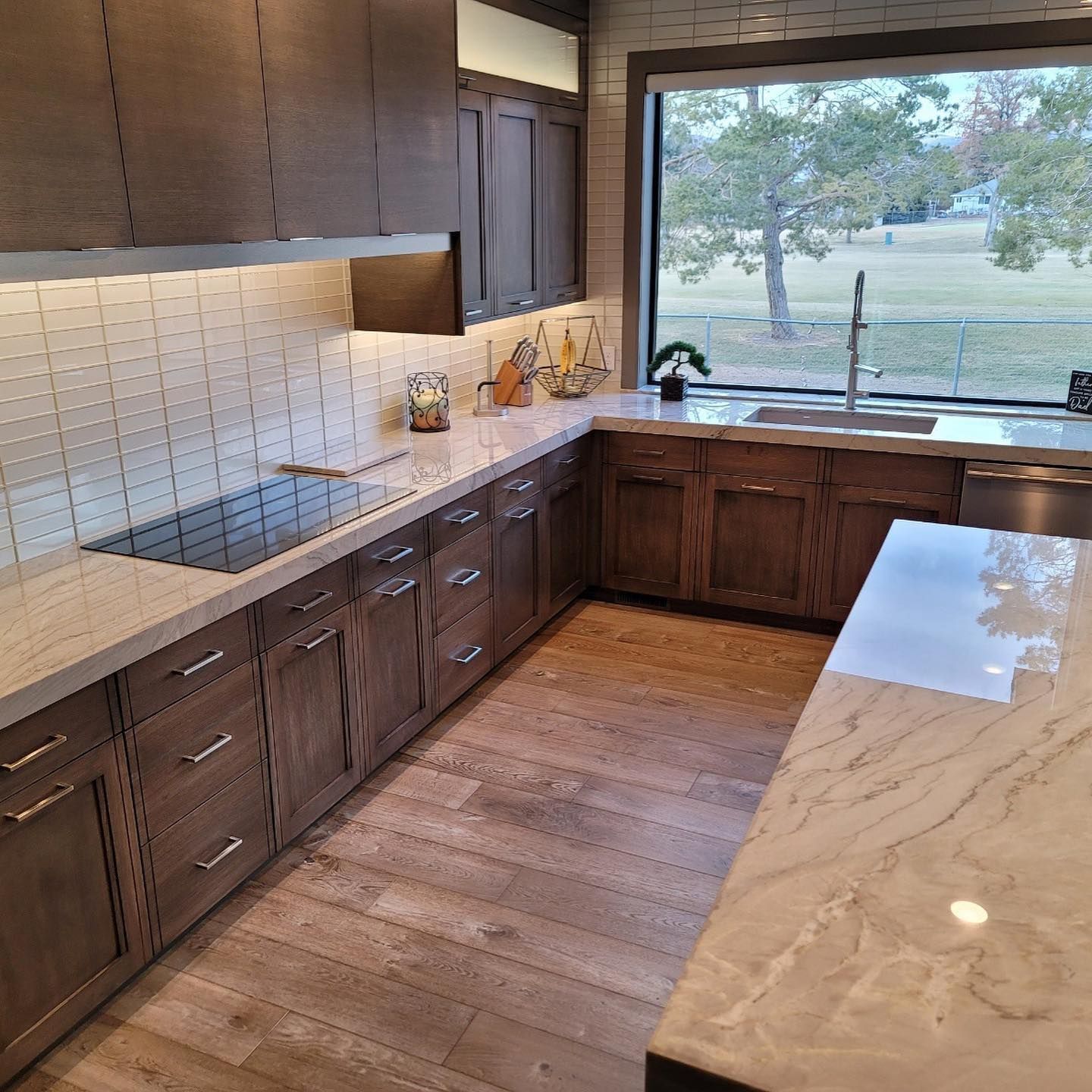 Modern kitchen with wood cabinets, white countertops, and a large window overlooking a yard.